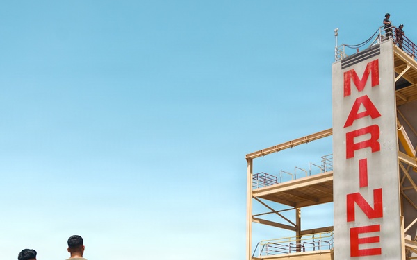 A group of civilians and U.S. Marines gather at the base of a tall training tower marked with the word 'MARINES' in large red letters on a sandy beach. The scene appears to be a public outreach or recruitment event, with military personnel in camouflage uniforms interacting with visitors. The setting is a sunny coastal location with a residential hillside visible in the background.