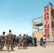 A group of civilians and U.S. Marines gather at the base of a tall training tower marked with the word 'MARINES' in large red letters on a sandy beach. The scene appears to be a public outreach or recruitment event, with military personnel in camouflage uniforms interacting with visitors. The setting is a sunny coastal location with a residential hillside visible in the background.