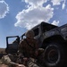 A U.S. Army soldier in full combat gear and helmet is performing tactical combat casualty care (TCCC) on a fellow soldier lying on the ground near a military HMMWV (Humvee). The setting appears to be an arid, desert training environment with red dirt terrain. The vehicle has markings consistent with a training or exercise scenario, including chalked text on its side.