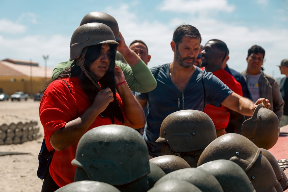 A group of civilians are trying on military-style steel helmets, likely during a base tour or public outreach event. A woman in a red shirt is adjusting a helmet on her head in the foreground, while several other participants stand behind a table stacked with additional helmets. The setting appears to be an outdoor military or training facility under bright daylight conditions.