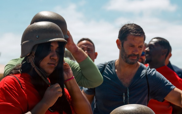 A group of civilians are trying on military-style steel helmets, likely during a base tour or public outreach event. A woman in a red shirt is adjusting a helmet on her head in the foreground, while several other participants stand behind a table stacked with additional helmets. The setting appears to be an outdoor military or training facility under bright daylight conditions.