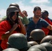 A group of civilians are trying on military-style steel helmets, likely during a base tour or public outreach event. A woman in a red shirt is adjusting a helmet on her head in the foreground, while several other participants stand behind a table stacked with additional helmets. The setting appears to be an outdoor military or training facility under bright daylight conditions.