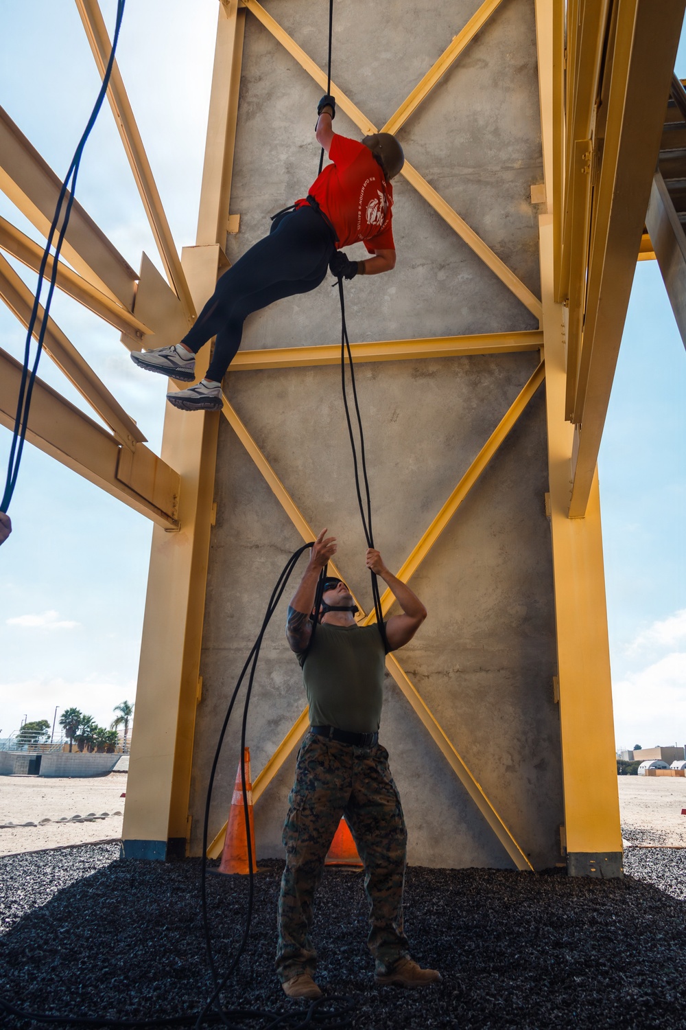 A person in a red shirt and black pants is rappelling down a concrete training tower with yellow steel framework, while a Marine in camouflage utilities and olive drab t-shirt acts as a belayer on the ground controlling the rope. The setting appears to be a military installation rappelling tower, likely used for training exercises. The Marine is managing the safety rope from below while the rappeller descends the wall face.