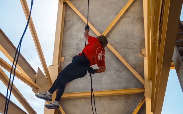A person in a red shirt and black pants is rappelling down a concrete training tower with yellow steel framework, while a Marine in camouflage utilities and olive drab t-shirt acts as a belayer on the ground controlling the rope. The setting appears to be a military installation rappelling tower, likely used for training exercises. The Marine is managing the safety rope from below while the rappeller descends the wall face.