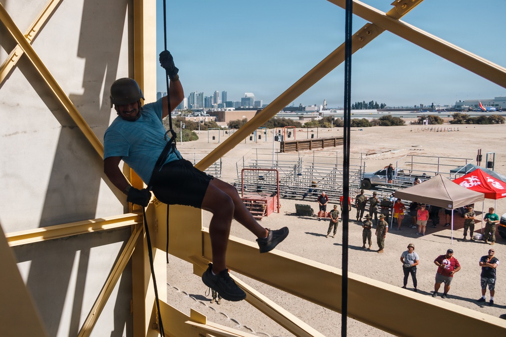 A person in civilian clothing is rappelling down a yellow steel training tower at what appears to be a military installation, likely a Marine Corps base given the visible USMC red flag/tent in the background. Spectators and uniformed military personnel observe from the ground below. A city skyline is visible in the distance, consistent with the San Diego area.