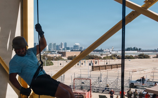 A person in civilian clothing is rappelling down a yellow steel training tower at what appears to be a military installation, likely a Marine Corps base given the visible USMC red flag/tent in the background. Spectators and uniformed military personnel observe from the ground below. A city skyline is visible in the distance, consistent with the San Diego area.