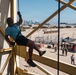 A person in civilian clothing is rappelling down a yellow steel training tower at what appears to be a military installation, likely a Marine Corps base given the visible USMC red flag/tent in the background. Spectators and uniformed military personnel observe from the ground below. A city skyline is visible in the distance, consistent with the San Diego area.