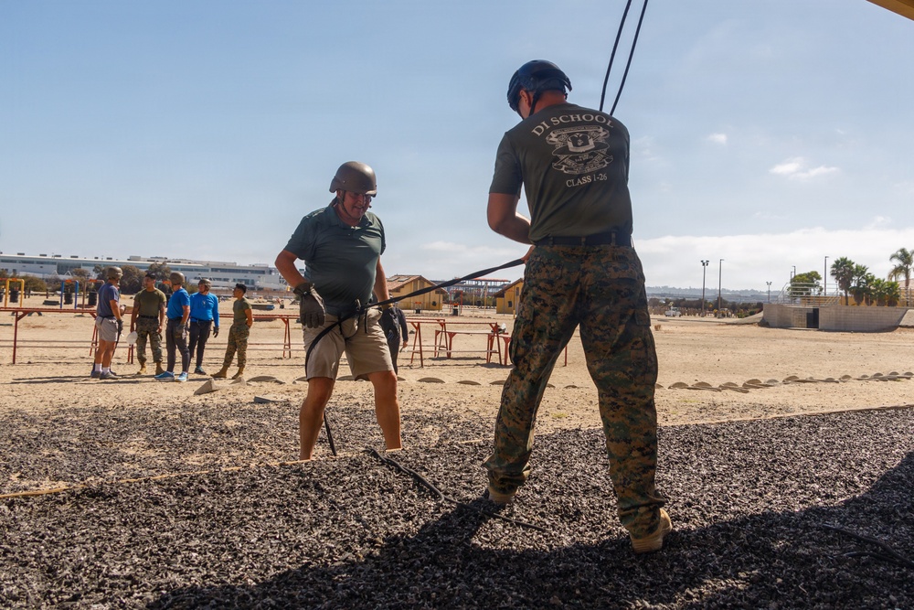 A military training exercise is taking place at what appears to be a Marine Corps base, likely MCRD San Diego. A civilian or visitor wearing a helmet and harness is being assisted by a Marine in MARPAT camouflage uniform from 'DI School Class I-26' during what appears to be a rappelling or confidence course activity. A group of observers, including civilians in casual clothing and military personnel, watch from the background.
