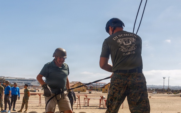 A military training exercise is taking place at what appears to be a Marine Corps base, likely MCRD San Diego. A civilian or visitor wearing a helmet and harness is being assisted by a Marine in MARPAT camouflage uniform from 'DI School Class I-26' during what appears to be a rappelling or confidence course activity. A group of observers, including civilians in casual clothing and military personnel, watch from the background.