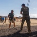 A military training exercise is taking place at what appears to be a Marine Corps base, likely MCRD San Diego. A civilian or visitor wearing a helmet and harness is being assisted by a Marine in MARPAT camouflage uniform from 'DI School Class I-26' during what appears to be a rappelling or confidence course activity. A group of observers, including civilians in casual clothing and military personnel, watch from the background.