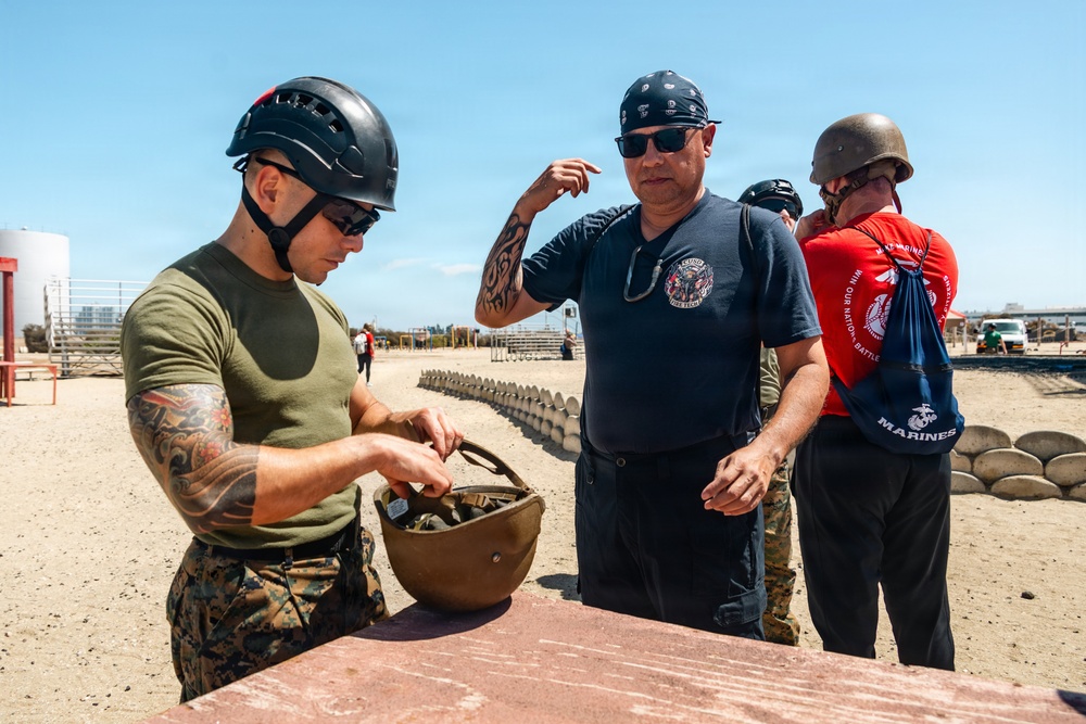 A U.S. Marine in uniform and tactical helmet is demonstrating or explaining a military helmet to a civilian visitor wearing a fire department t-shirt. The scene appears to take place at a military training facility or base, likely during a public outreach or familiarization event. A third person in a red Marines t-shirt and WWII-style helmet is visible in the background.