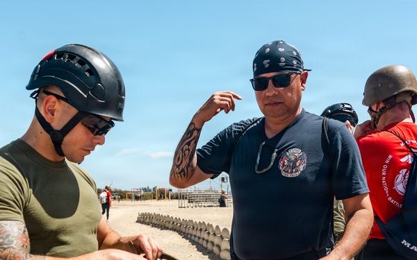 A U.S. Marine in uniform and tactical helmet is demonstrating or explaining a military helmet to a civilian visitor wearing a fire department t-shirt. The scene appears to take place at a military training facility or base, likely during a public outreach or familiarization event. A third person in a red Marines t-shirt and WWII-style helmet is visible in the background.