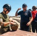 A U.S. Marine in uniform and tactical helmet is demonstrating or explaining a military helmet to a civilian visitor wearing a fire department t-shirt. The scene appears to take place at a military training facility or base, likely during a public outreach or familiarization event. A third person in a red Marines t-shirt and WWII-style helmet is visible in the background.