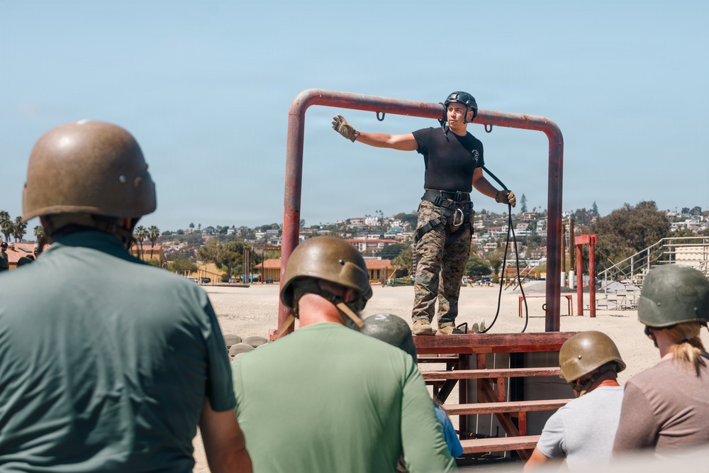 A military instructor in camouflage pants and black shirt stands on an elevated red metal platform, demonstrating rappelling or rope techniques to a group of observers wearing helmets. The setting appears to be an outdoor military training facility, likely Marine Corps Recruit Depot (MCRD) San Diego based on the visible terrain and architecture. The instructor is equipped with a harness and rope, gesturing with one arm extended while addressing the group below.