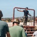 A military instructor in camouflage pants and black shirt stands on an elevated red metal platform, demonstrating rappelling or rope techniques to a group of observers wearing helmets. The setting appears to be an outdoor military training facility, likely Marine Corps Recruit Depot (MCRD) San Diego based on the visible terrain and architecture. The instructor is equipped with a harness and rope, gesturing with one arm extended while addressing the group below.