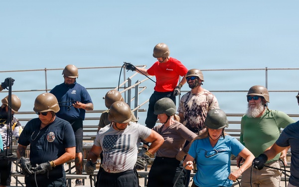 A group of approximately 12-15 people wearing olive/tan military-style helmets are standing in front of bleachers on a sandy beach area, handling ropes or cables. The setting appears to be an outdoor training exercise or team-building event, with participants dressed in casual civilian clothing. One person in a red shirt is elevated on the bleachers, appearing to assist or instruct the group.