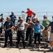 A group of approximately 12-15 people wearing olive/tan military-style helmets are standing in front of bleachers on a sandy beach area, handling ropes or cables. The setting appears to be an outdoor training exercise or team-building event, with participants dressed in casual civilian clothing. One person in a red shirt is elevated on the bleachers, appearing to assist or instruct the group.