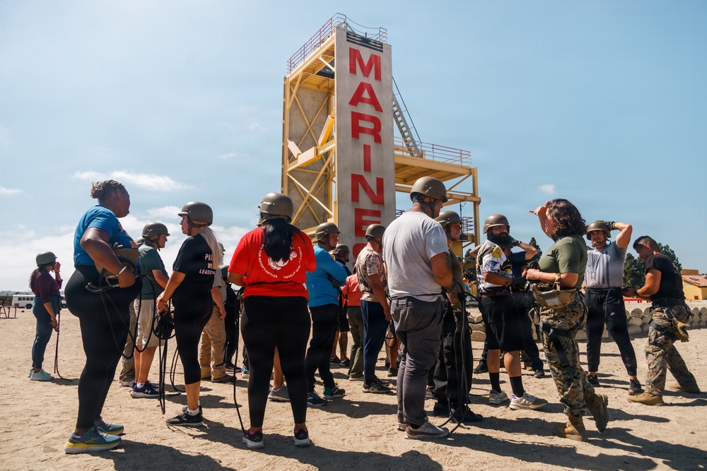 A group of civilians wearing military-style helmets and harnesses are gathered on a sandy training ground at what appears to be a U.S. Marine Corps base. They are standing in front of a tall training tower with 'MARINES' written in large red letters. A Marine Corps instructor in uniform appears to be briefing or directing the group, suggesting this is a civilian orientation or community outreach event.