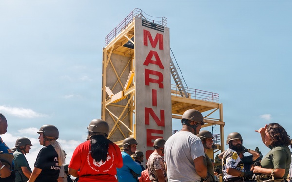 A group of civilians wearing military-style helmets and harnesses are gathered on a sandy training ground at what appears to be a U.S. Marine Corps base. They are standing in front of a tall training tower with 'MARINES' written in large red letters. A Marine Corps instructor in uniform appears to be briefing or directing the group, suggesting this is a civilian orientation or community outreach event.