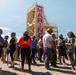 A group of civilians wearing military-style helmets and harnesses are gathered on a sandy training ground at what appears to be a U.S. Marine Corps base. They are standing in front of a tall training tower with 'MARINES' written in large red letters. A Marine Corps instructor in uniform appears to be briefing or directing the group, suggesting this is a civilian orientation or community outreach event.
