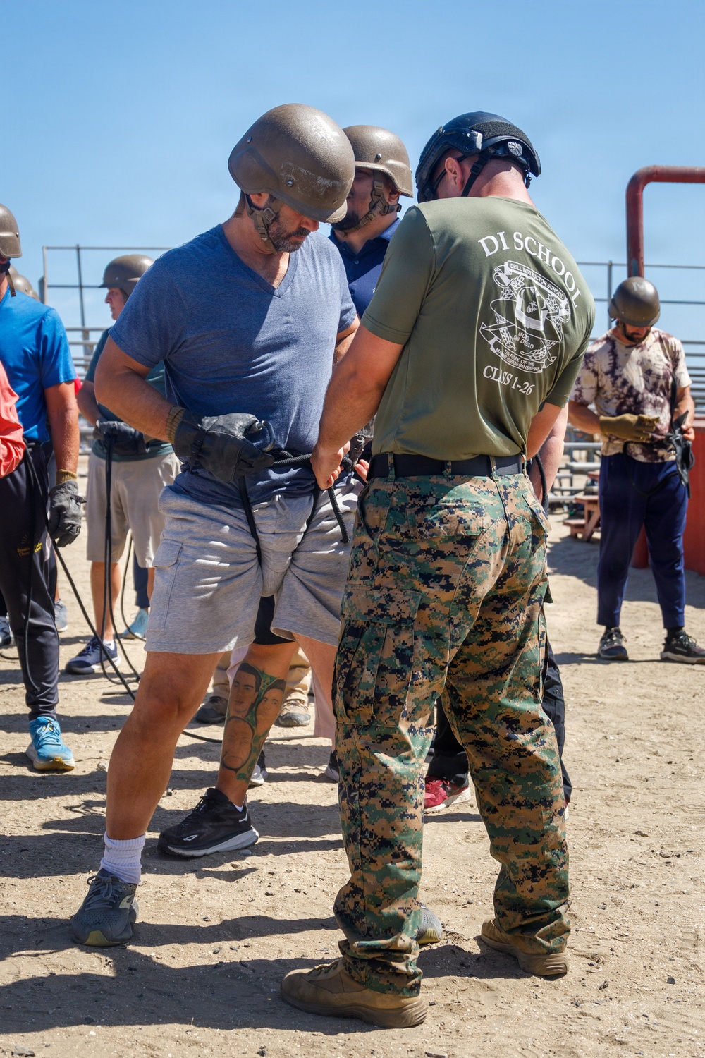 A Marine Corps Drill Instructor School instructor in MARPAT camouflage and a 'DI SCHOOL CLASS 1-26' t-shirt is assisting a civilian participant wearing a tactical helmet and gloves during what appears to be a military familiarization or media/VIP experience event. Multiple civilians in casual clothing are wearing military-style helmets and participating in various activities on a sandy outdoor training area. The setting appears to be a military base or training facility.