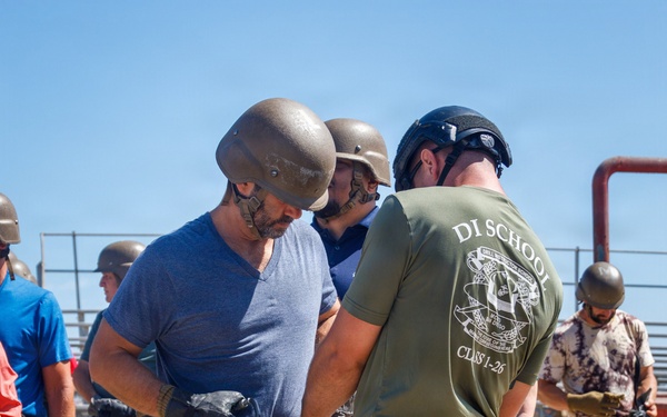 A Marine Corps Drill Instructor School instructor in MARPAT camouflage and a 'DI SCHOOL CLASS 1-26' t-shirt is assisting a civilian participant wearing a tactical helmet and gloves during what appears to be a military familiarization or media/VIP experience event. Multiple civilians in casual clothing are wearing military-style helmets and participating in various activities on a sandy outdoor training area. The setting appears to be a military base or training facility.