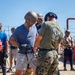 A Marine Corps Drill Instructor School instructor in MARPAT camouflage and a 'DI SCHOOL CLASS 1-26' t-shirt is assisting a civilian participant wearing a tactical helmet and gloves during what appears to be a military familiarization or media/VIP experience event. Multiple civilians in casual clothing are wearing military-style helmets and participating in various activities on a sandy outdoor training area. The setting appears to be a military base or training facility.