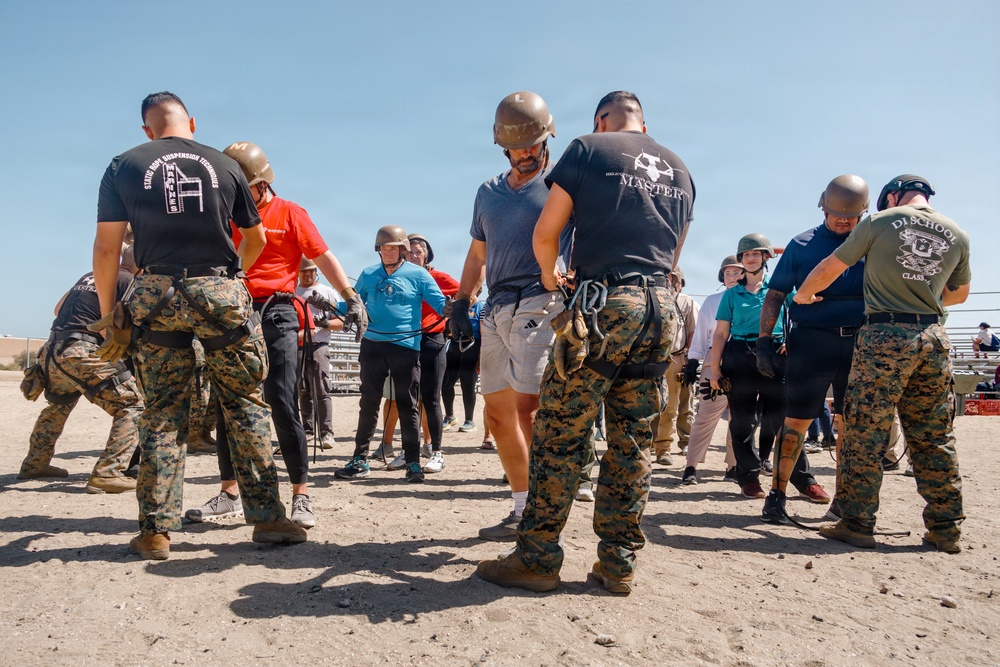 A group of civilians and military personnel in MARPAT camouflage uniforms are engaged in what appears to be a training or orientation exercise on a sandy outdoor area. Several participants are wearing military-style helmets, and instructors in black shirts with military insignia are guiding the group. The setting and attire suggest a military familiarization or civilian-military engagement event.