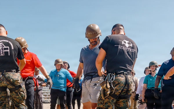 A group of civilians and military personnel in MARPAT camouflage uniforms are engaged in what appears to be a training or orientation exercise on a sandy outdoor area. Several participants are wearing military-style helmets, and instructors in black shirts with military insignia are guiding the group. The setting and attire suggest a military familiarization or civilian-military engagement event.