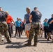 A group of civilians and military personnel in MARPAT camouflage uniforms are engaged in what appears to be a training or orientation exercise on a sandy outdoor area. Several participants are wearing military-style helmets, and instructors in black shirts with military insignia are guiding the group. The setting and attire suggest a military familiarization or civilian-military engagement event.