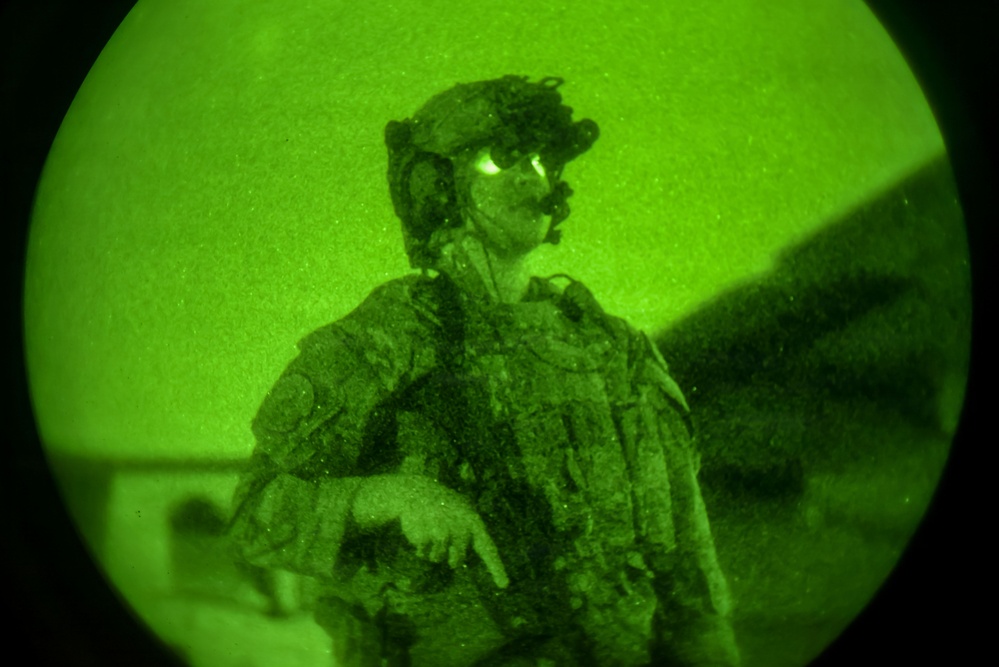 A night vision image captured through a circular lens showing a military personnel in full tactical gear. The green monochromatic image is characteristic of night vision optics (NVG - Night Vision Goggles) photography. The subject appears to be in an operational environment, possibly near a vehicle or aircraft.