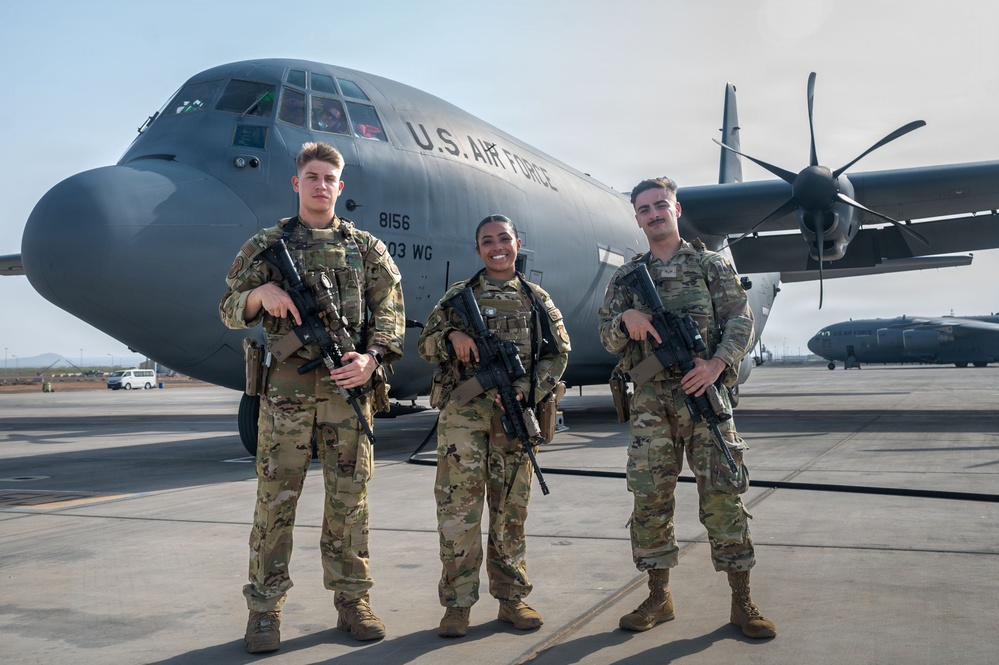 Three U.S. Air Force personnel in MultiCam/OCP camouflage uniforms are posed on a flight line in front of a C-130 Hercules transport aircraft marked 'U.S. AIR FORCE' with tail number 8156 and unit designation '03 WG'. All three individuals are holding M4-style carbine rifles and appear to be in a formal posed photograph. A second aircraft is visible in the background on the right side of the frame.