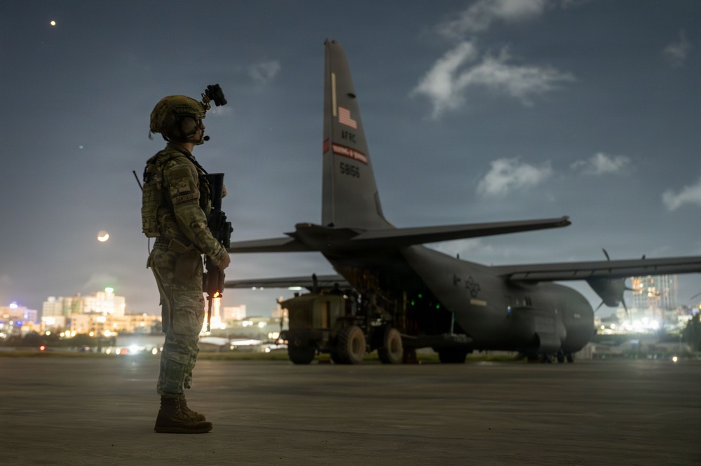 A fully equipped military service member stands guard on a tarmac at night beside a U.S. Air Force C-130 Hercules transport aircraft. The soldier is wearing combat gear including a helmet with night-vision device mount, body armor, and carrying a rifle. City lights are visible in the background, suggesting proximity to an urban area.