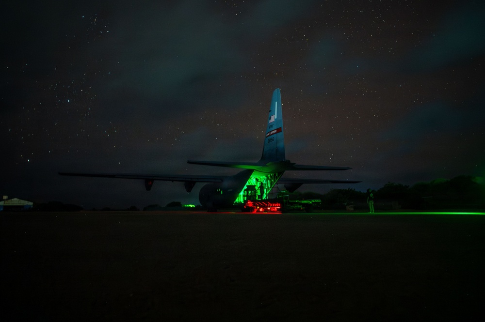 A military transport aircraft, consistent with a C-130 Hercules, is photographed at night on a tarmac under a star-filled sky. The aircraft's rear cargo ramp is open and illuminated by bright green interior lighting, with red lighting visible beneath what appears to be ground support equipment. A lone figure stands to the right of the aircraft, and a green laser or light beam extends horizontally across the tarmac.