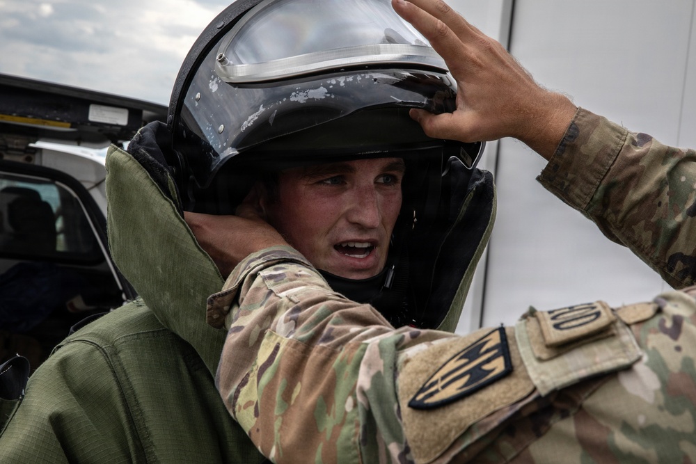 A military personnel wearing a protective bomb disposal or EOD (Explosive Ordnance Disposal) suit and helmet is being assisted by another soldier in multicam/OCP camouflage uniform. The soldier assisting appears to be adjusting or securing the helmet visor of the person in the protective suit. The setting appears to be an outdoor military or training environment near vehicles.
