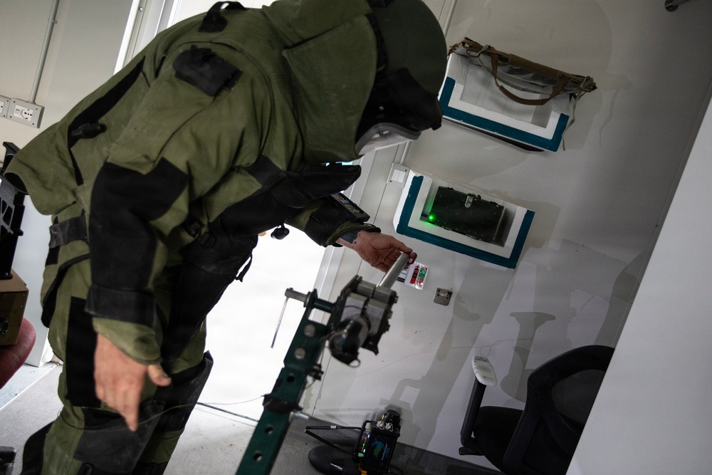 A person wearing a green EOD (Explosive Ordnance Disposal) bomb suit is leaning forward and examining or interacting with a wall-mounted panel or device inside what appears to be a training facility or mobile unit. The individual is wearing full protective gear including a helmet and armored suit. A green indicator light is visible on a framed panel on the wall, and robotic or mechanical equipment is visible in the foreground.