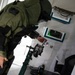 A person wearing a green EOD (Explosive Ordnance Disposal) bomb suit is leaning forward and examining or interacting with a wall-mounted panel or device inside what appears to be a training facility or mobile unit. The individual is wearing full protective gear including a helmet and armored suit. A green indicator light is visible on a framed panel on the wall, and robotic or mechanical equipment is visible in the foreground.
