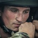 A close-up portrait of a young male soldier wearing a military helmet and multicam/camouflage uniform. His fist is raised near his chin, and he appears focused or in a moment of concentration or fatigue. The image is a high-quality, dramatic military portrait with shallow depth of field.