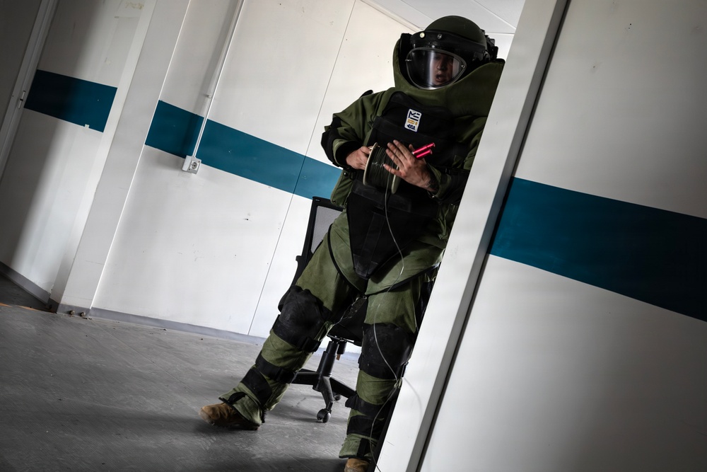 A person wearing a full EOD (Explosive Ordnance Disposal) bomb suit is navigating through a doorway inside what appears to be a building used for training exercises. The suit is olive green with black protective panels, and the individual is holding a red cylindrical object while carefully moving through the entrance. The setting appears to be an indoor training facility with white walls and a teal/blue horizontal stripe.