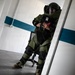 A person wearing a full EOD (Explosive Ordnance Disposal) bomb suit is navigating through a doorway inside what appears to be a building used for training exercises. The suit is olive green with black protective panels, and the individual is holding a red cylindrical object while carefully moving through the entrance. The setting appears to be an indoor training facility with white walls and a teal/blue horizontal stripe.