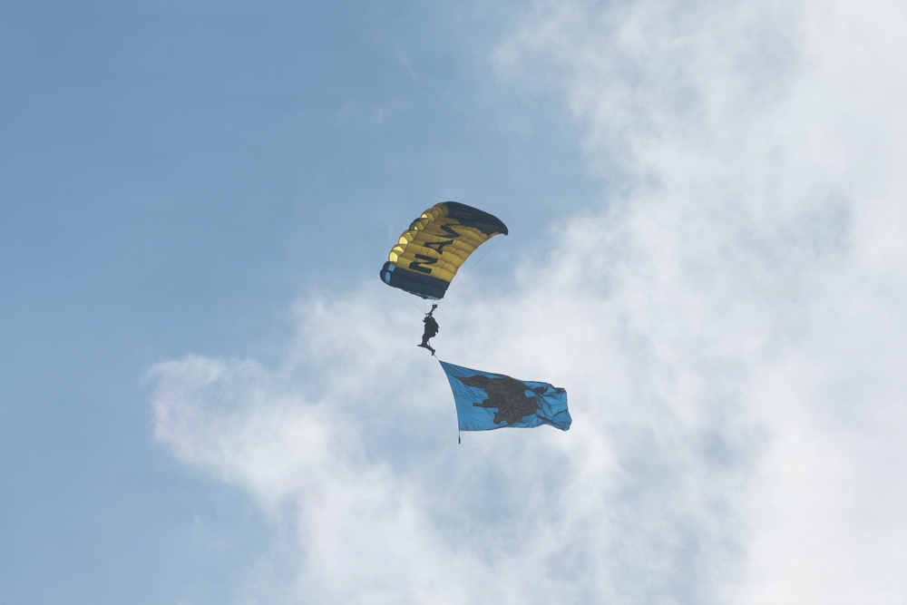 A parachutist is descending through a partly cloudy blue sky using a yellow and black canopy with 'NAVY' written on it. The jumper is trailing a light blue flag featuring a dark silhouette emblem, consistent with a U.S. Navy parachute demonstration team performance. The image appears to be taken from the ground looking upward.