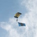 A parachutist is descending through a partly cloudy blue sky using a yellow and black canopy with 'NAVY' written on it. The jumper is trailing a light blue flag featuring a dark silhouette emblem, consistent with a U.S. Navy parachute demonstration team performance. The image appears to be taken from the ground looking upward.