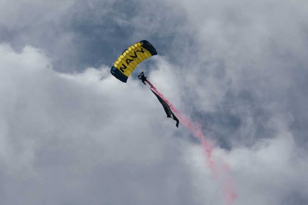 A U.S. Navy parachutist is descending through a cloudy sky with a yellow and black canopy bearing the word 'NAVY'. The jumper is trailing pink/red colored smoke, indicative of a demonstration or airshow performance. The overcast sky provides a dramatic backdrop to the display.