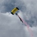A U.S. Navy parachutist is descending through a cloudy sky with a yellow and black canopy bearing the word 'NAVY'. The jumper is trailing pink/red colored smoke, indicative of a demonstration or airshow performance. The overcast sky provides a dramatic backdrop to the display.