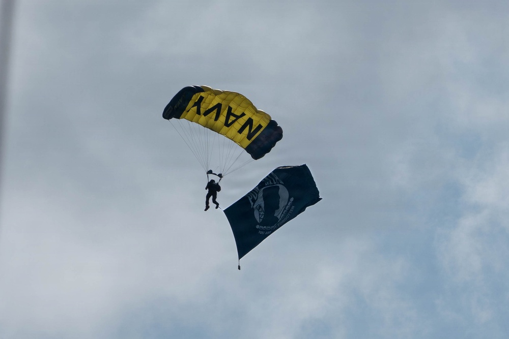 A military skydiver descends under a yellow and black parachute bearing the word 'NAVY' against an overcast sky. The jumper is carrying a large dark flag that appears to be a POW/MIA flag, identifiable by the silhouetted head and surrounding text. This appears to be a ceremonial military demonstration jump, likely at a public event or airshow.