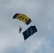 A military skydiver descends under a yellow and black parachute bearing the word 'NAVY' against an overcast sky. The jumper is carrying a large dark flag that appears to be a POW/MIA flag, identifiable by the silhouetted head and surrounding text. This appears to be a ceremonial military demonstration jump, likely at a public event or airshow.