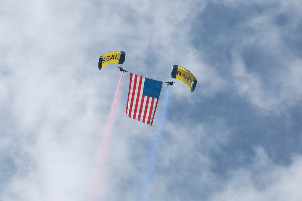 Two skydivers with yellow and black parachutes are descending while carrying a large American flag between them. The parachutes are labeled 'SEAL' and 'NAVY', identifying the jumpers as U.S. Navy SEALs performing a demonstration. Red and blue smoke trails stream downward from the jumpers against a partly cloudy sky.