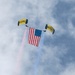 Two skydivers with yellow and black parachutes are descending while carrying a large American flag between them. The parachutes are labeled 'SEAL' and 'NAVY', identifying the jumpers as U.S. Navy SEALs performing a demonstration. Red and blue smoke trails stream downward from the jumpers against a partly cloudy sky.