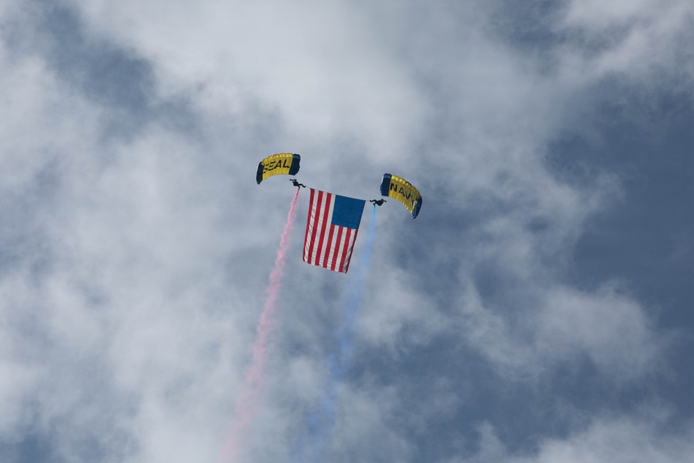 Two military skydivers are descending with yellow parachutes labeled 'SEAL' and 'NAVY', carrying a large American flag between them. Red and blue smoke trails stream from the jumpers, creating a patriotic aerial display against a partly cloudy sky. This appears to be a demonstration by the U.S. Navy SEAL parachute team at a public event or air show.