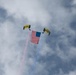 Two military skydivers are descending with yellow parachutes labeled 'SEAL' and 'NAVY', carrying a large American flag between them. Red and blue smoke trails stream from the jumpers, creating a patriotic aerial display against a partly cloudy sky. This appears to be a demonstration by the U.S. Navy SEAL parachute team at a public event or air show.